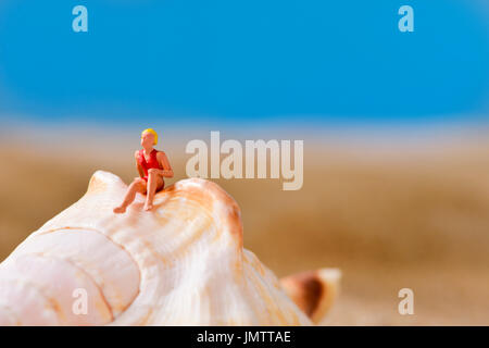 Nahaufnahme einer Miniatur-Frau mit Badeanzug entspannt auf einer Muschel am Strand, mit einem negativen Raum an der Spitze Stockfoto