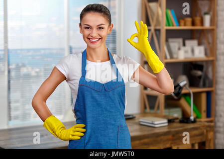 Studioaufnahme der Haushälterin. Schöne Frau Reinigung Büro. Frau mit Handschuhen, lächelnd und zeigt ok Sign. Stockfoto