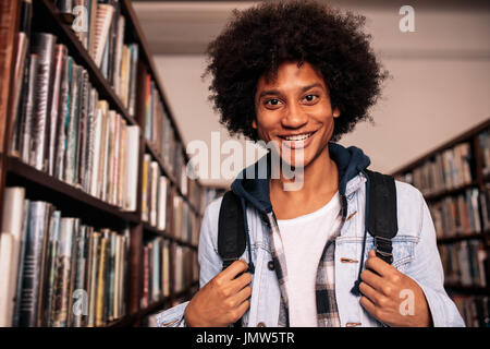 Student in der Bibliothek. Junger afrikanischer Mann lächelt und blickt mit Rucksack in die Kamera. Stockfoto