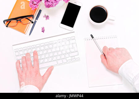 Mann schreiben Notizen im Tagebuch. Arbeitsplatz mit Computer-Tastatur, Handy, Tasse Kaffee, Notizbuch, Kugelschreiber, Bleistift, Gläser und lila Blumen auf der whi Stockfoto