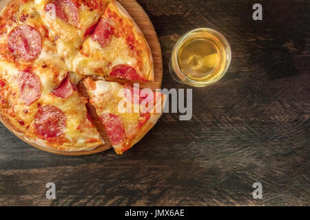 Eine obenliegende Foto eine Peperoni-Pizza mit einer Scheibe abschneiden, mit einem Glas Weißwein ablöschen, geschossen von oben auf eine dunkle rustikale Textur mit einem Platz für tex Stockfoto