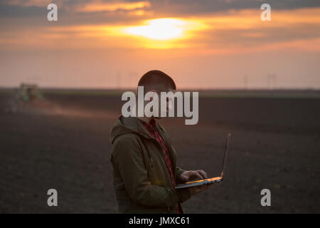 Junglandwirt mit Laptop stand im Feld mit Traktor Eggen im Hintergrund Stockfoto