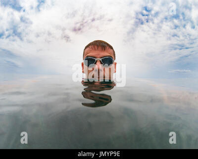 Ernste Jüngling im Meer, sein Kopf ist auf der glatten Oberfläche des Wassers, über ihn den bewölkten Himmel, er trägt Brille Stockfoto