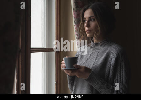 Nahaufnahme der nachdenkliche Frau mit Kaffee im stehen durch die Fenster zu Hause Stockfoto
