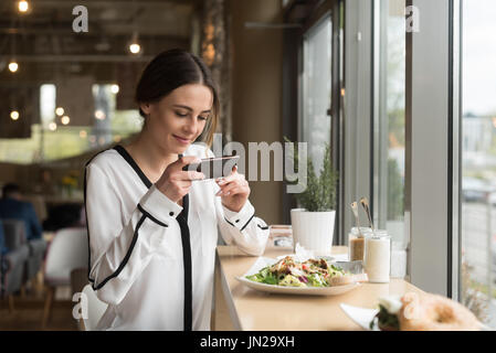 Lächelnde Frau fotografieren Essen im Teller am Tisch im café Stockfoto