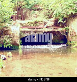 Kleinen Wehr am Fluss fließt aus der Höhle. Kaltes Wasser des kleinen Fluss über kleine steinerne Wehr.  Steinige und rostigen Konstruktion. Stockfoto