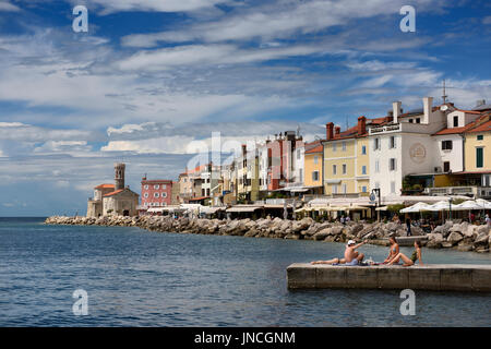 Junge Leute, Sonnenbaden am Dock in Piran Slowenien an der Adria-Küste mit der 13. Jahrhundert Kirche von St Clement am Leuchtturm von Punta Stockfoto