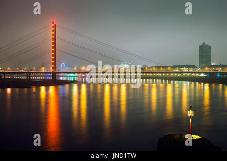 Brücke über den Rhein in Düsseldorf-Stadt in der Nacht mit bunten Beleuchtung spiegelt sich im Wasser. Einsame Laterne am Ufer des Flusses. Stockfoto