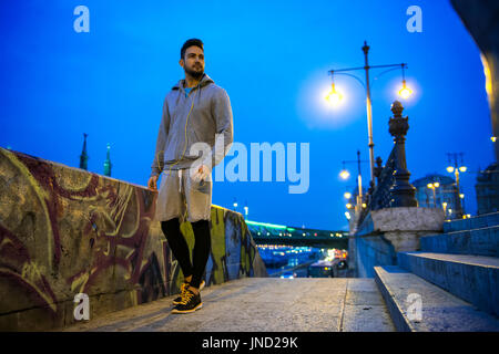 Ein hübscher Junge Sportler in die Lichter der Stadt am Flussufer in der Nacht mit einer Brücke hinter ihm stehen Stockfoto