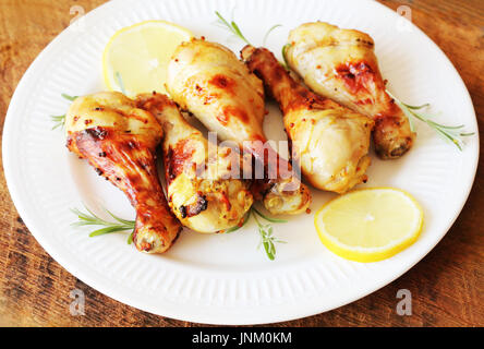 Gegrillte Hähnchenschenkel mit Senf auf Holztisch auf weißen Teller mit Rosmarin und Zitrone serviert. BBQ-Abendessen-Hintergrund Stockfoto