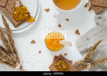 Süße Snacks und Frühstück mit Roggen Knäckebrot (schwedische Kekse), Ausbreitung orange Marmelade, Tasse mit grünem Tee, Marmelade im Glas jar, getrocknete Blumen, auf weißen bac Stockfoto