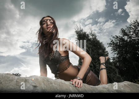 Junge schöne Frau, gekleidet in ein Lederbekleidung sah aus wie ein Amazon oder Krieger liegen auf einem großen Stein. Dramatische Beautyshot Stockfoto