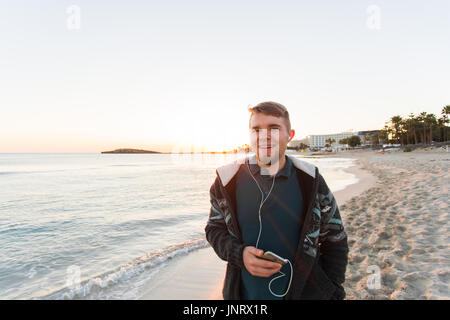 Stilvolle junger Mann hören Sie Musik im Kopfhörer auf einem Smartphone am Strand. Lifestyle, Technik und Menschen-Konzept Stockfoto