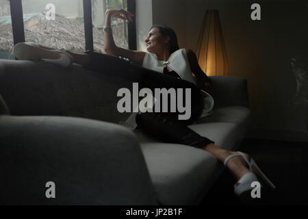 Schöne Frau, die durch große helle Fenster am Wohngebäude liegend in großen bequemen Sessel. Stockfoto