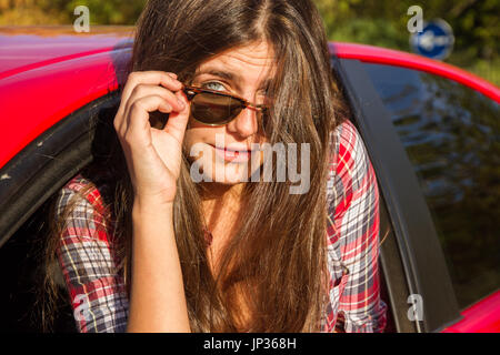 Porträt der glückliche junge Frau, die auf einem Road Trip lehnte sich aus dem Fenster des roten Auto. Sie hat blaue Augen und braune Haare. Stockfoto
