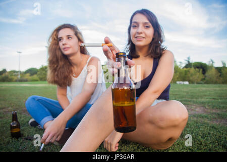 Zwei Mädchen, Freunde, Zigaretten rauchen und trinken Bier Flaschen in den Park an einem bewölkten Tag. Sie sind von der Feier und sind glücklich. Stockfoto