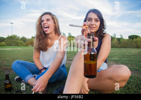 Zwei Mädchen, Freunde, Zigaretten rauchen und trinken Bier Flaschen in den Park an einem bewölkten Tag. Sie sind von der Feier und sind glücklich. Stockfoto