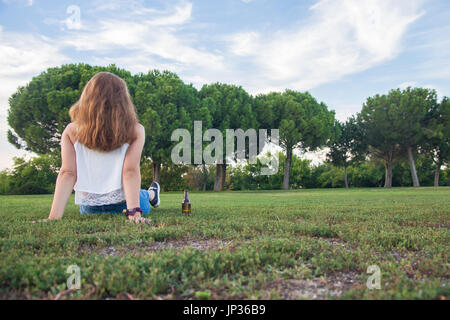 Mädchen hat ihre Bierflasche auf dem Boden, während am Horizont nachdenklich. Ihre Hand und Bier ruht auf dem Gras. Stockfoto