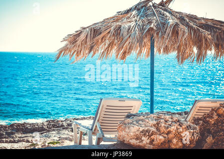 Felsiger Strand in der Nähe von azurblauem Meer Strand zwei Liegestühlen unter Dach. Urlaub Ferien Hintergrundbild Stockfoto