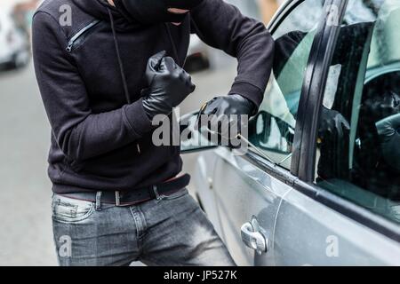 Der Mann gekleidet in schwarz mit einer Sturmhaube auf seinem Kopf versucht, ins Auto zu brechen. Er nutzt einen Schraubenzieher. Autodieb, Auto-Diebstahl-Konzept Stockfoto