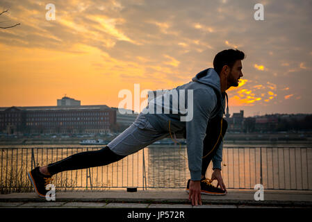 Ein hübscher junger Mann stretching und Aufwärmen am Flussufer mit den Sonnenuntergang hinter ihm Stockfoto