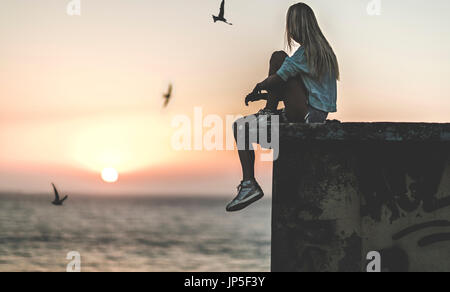 Seitenansicht einer jungen Frau sitzt auf einer Mauer mit einem Sonnenuntergang hinter. Stockfoto