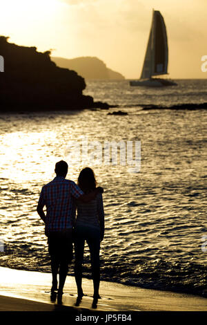 Rückansicht des paar stehend auf einem sandigen Strand durch das Meer, Klippen und ein Segelboot in der Ferne bei Sonnenuntergang. Stockfoto