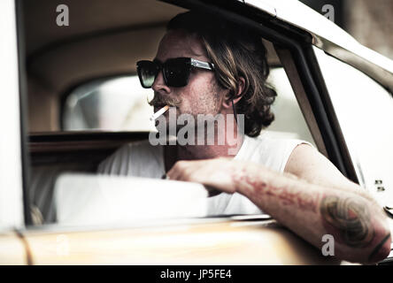Mann mit Schnurrbart, sitzen im Auto, das Tragen von Sonnenbrillen, rauchte eine Zigarette, tätowierten Arm schiefen aus Fenster. Stockfoto