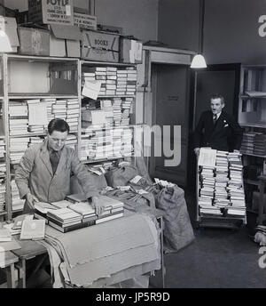 der 1950er Jahre, Bild zeigt einen beschichteten Mitarbeiter mit Krawatte Verpackung Bücher bereit für den Versand in eine Abstellkammer in der berühmten Buchhandlung W & G Foyle Ltd, Charing Cross Road, London, während ein anderer geeignet Mitarbeiter mehr bringt Bücher, auf einem Wagen. Stockfoto