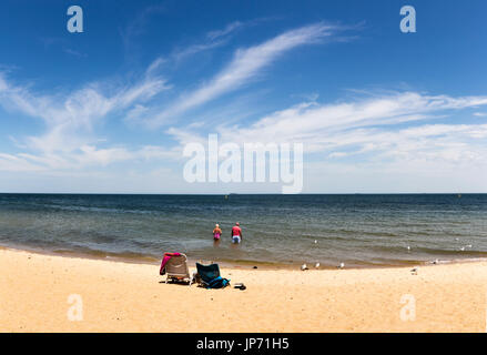 Melbourne, Victoria, Australien. Älteres paar waten und entfernten Schiffe mit Liegestühlen am Strand zu beobachten. Panorama. Stockfoto