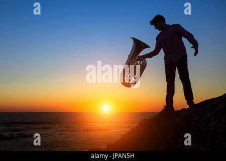 Silhouette des Musikers mit der Trompete auf felsigen Küste bei traumhaften Sonnenuntergang. Tuba-Instrument. Stockfoto