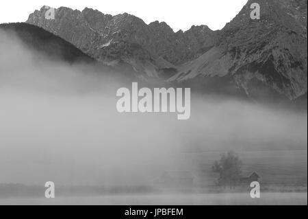 Neblige Stimmung am Fuße des Karwendelgebirges (Berge) s. Die Ufer des Schmalensee (See) mit zwei Heustadel im Vordergrund. Stockfoto