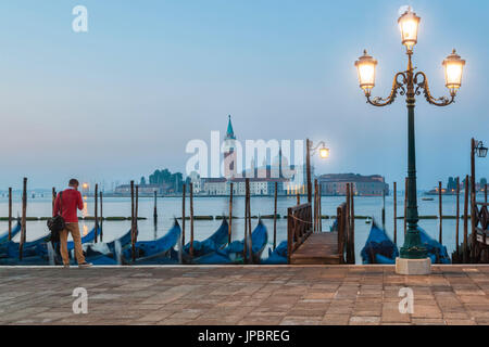 Ein einsamer Mann wartet vor der Gondeln, die entlang der Riva Degli Schiavoni angedockt. im Hintergrund die Insel San Giorgio Maggiore, Venedig, Italien Stockfoto