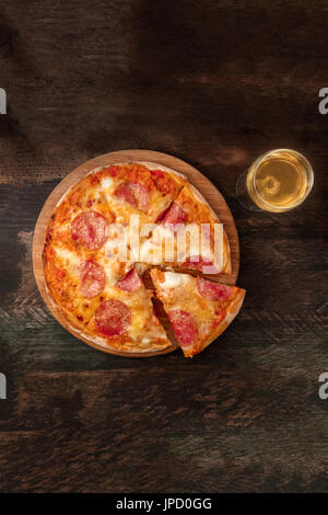 Eine obenliegende Foto eine Peperoni-Pizza mit einer Scheibe abschneiden, mit einem Glas Weißwein ablöschen, geschossen von oben auf eine dunkle rustikale Textur mit einem Platz für tex Stockfoto