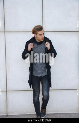 Schöne moderne blond Jungunternehmer mit blauen Augen posiert an eine Wand gelehnt trägt einen Mantel. Stockfoto