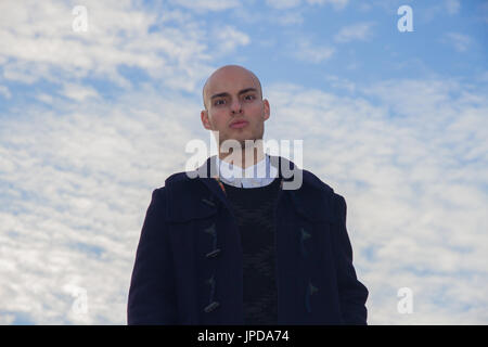 Kahl, junger Mann mit braunen Augen, gekleidet in moderne und stilvolle Kleidung auf Hintergrund blauer Himmel mit Wolken. Stockfoto