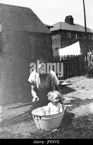 1930er Jahre, England, Badezeit, Mutter mit Baby-sitting in einem Blecheimer Baden außerhalb. Stockfoto