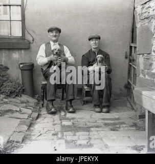 draußen Sie 1940er Jahre, England, Lancaster, zwei ältere nördliche Männer tragen Schiebermütze und Stiefel, in einem Hof auf kleine Stände mit Stolz ihre kleinen Hunde halten. Stockfoto