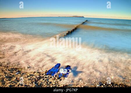 Maske mit blauen Flossen zum Schnorcheln bereit für den Einsatz auf steinigen Strand von hölzernen Wellenbrecher. Wellig Meeresspiegel. Stockfoto