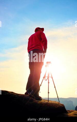 Fotograf mit Stativ auf Klippe und denken bleiben. Verträumte Kauz Landschaft, orange nebligen Sonnenaufgang in einem wunderschönen Tal unten Stockfoto
