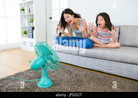 junge Mutter mit schönen kleinen Mädchen sitzen auf der Couch Sofa und machen Lachen posiert spielen elektrischen Lüfter kühlen Wind zu genießen. Stockfoto