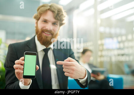 Porträt der schönen bärtigen Geschäftsmann präsentiert mobile app im Büro, auf Smartphone mit Greenscreen und glücklich lächelnd Stockfoto