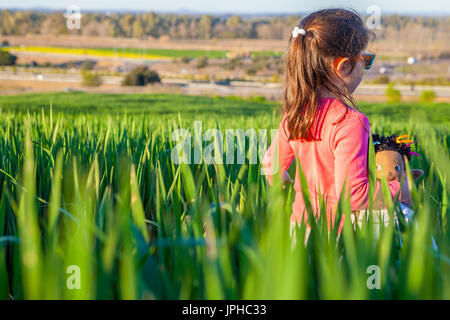 Kleine Mädchen und ihre Puppe zu Fuß durch grüne Getreide Feld bei Sonnenuntergang, Spanien Stockfoto