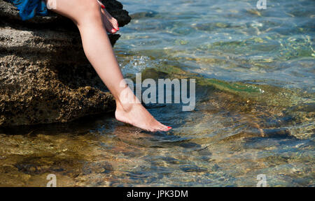 Junge Frau sitzt auf dem Stein legen seine Füße in sauberen und klaren Meerwasser. Sonniger Tag. Die horizontalen Rahmen. Stockfoto