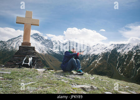 Frau ist Zeigefinger auf erstaunliche Berge Hintergrund, Laughing glückliche Wanderer Mädchen, Sonnenbrille und grauen Hut, blauer Jacken &amp; Mäntel Stockfoto