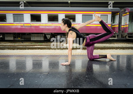Gesunde Frau, die Yoga macht, posiert draußen auf dem Boden eines Bahnhofs in Asien und trägt bunte Fitnesskleidung Stockfoto