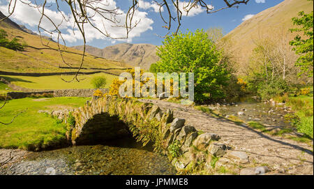 Eine atemberaubende Landschaft, alte Stein gewölbt Fußgängerbrücke über Tröpfelnden stream & Weg vorbei an Farmland zu baumlosen Hügel des Lake District Nat Pk Stockfoto