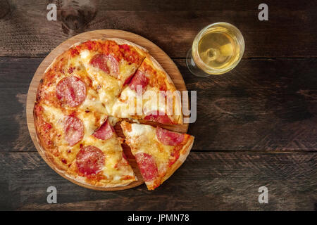 Eine obenliegende Foto eine Peperoni-Pizza mit einer Scheibe abschneiden, mit einem Glas Weißwein ablöschen, geschossen von oben auf eine dunkle rustikale Textur mit einem Platz für tex Stockfoto