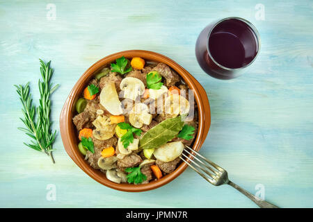 Ein Foto von einem Fleisch-Ragout in einer irdenen Schüssel mit einer Gabel und Zweige frischer Rosmarin, geschossen von oben auf eine Hintergrundtextur blaugrün, mit einem glas Stockfoto