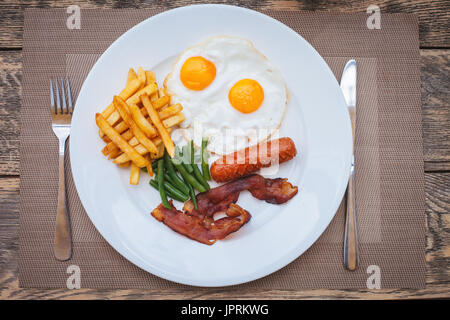 Englisches Frühstück mit Spiegeleiern, Speck, Würstchen, Bohnen und Kartoffeln Pommes Frites auf Holztisch Stockfoto